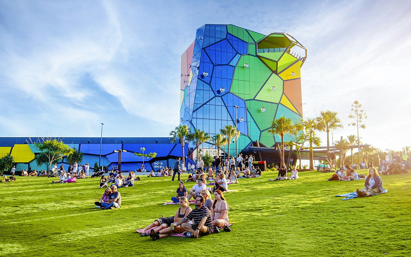 People relaxing on the lawn at Home of the Arts, Gold Coast, with colorful building in background.