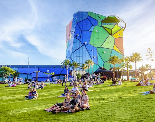 Persone che si rilassano sul prato dell'Home of the Arts, Gold Coast, con un edificio colorato sullo sfondo.