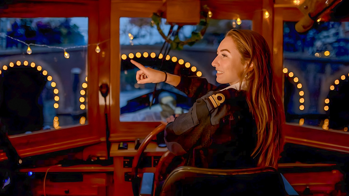 Skipper steering boat during Amsterdam Light Festival Cruise, pointing at illuminated canal bridge.