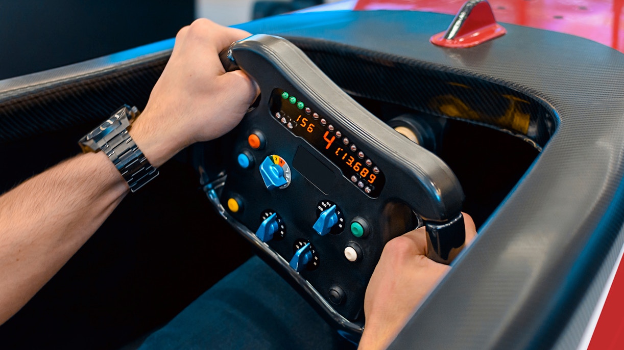 Hands gripping Ferrari F1 simulator steering wheel with digital display.