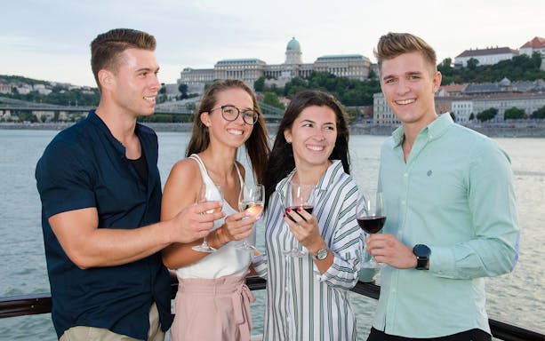 Group enjoying wine on Danube River sightseeing cruise with Buda Castle in background.