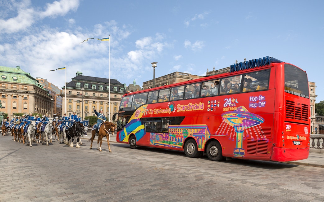 Red double-decker bus on a city sightseeing tour in Stockholm, passing a mounted royal guard parade.