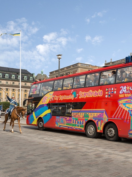 Red double-decker bus on a city sightseeing tour in Stockholm, passing a mounted royal guard parade.