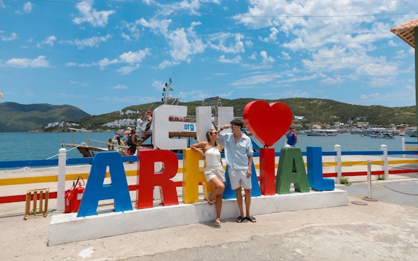 Tourists posing by colorful Arraial do Cabo sign with cruise boats in the background.