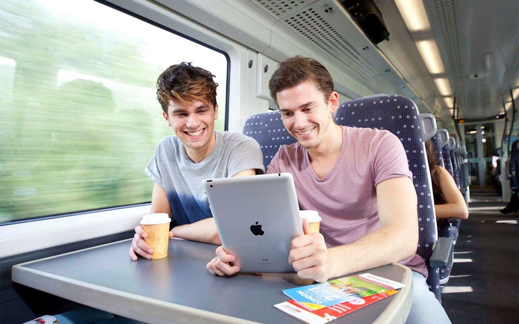 Two people enjoying a train ride with coffee and a tablet, brochures on the table.
