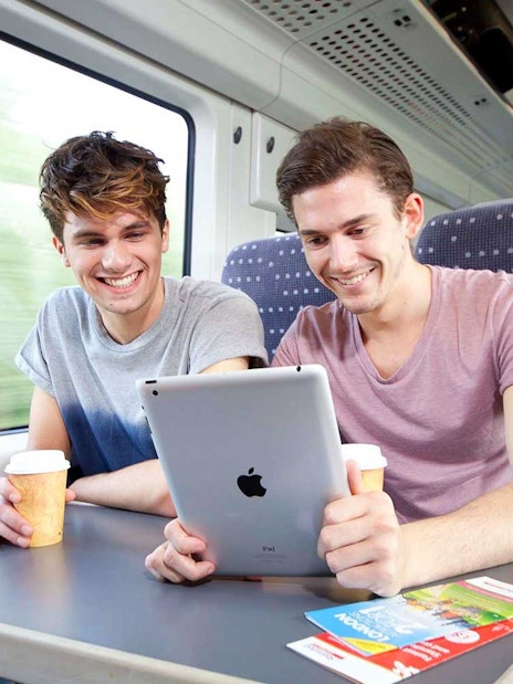 Two people enjoying a train ride with coffee and a tablet, brochures on the table.