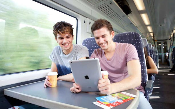 Two people enjoying a train ride with coffee and a tablet, brochures on the table.