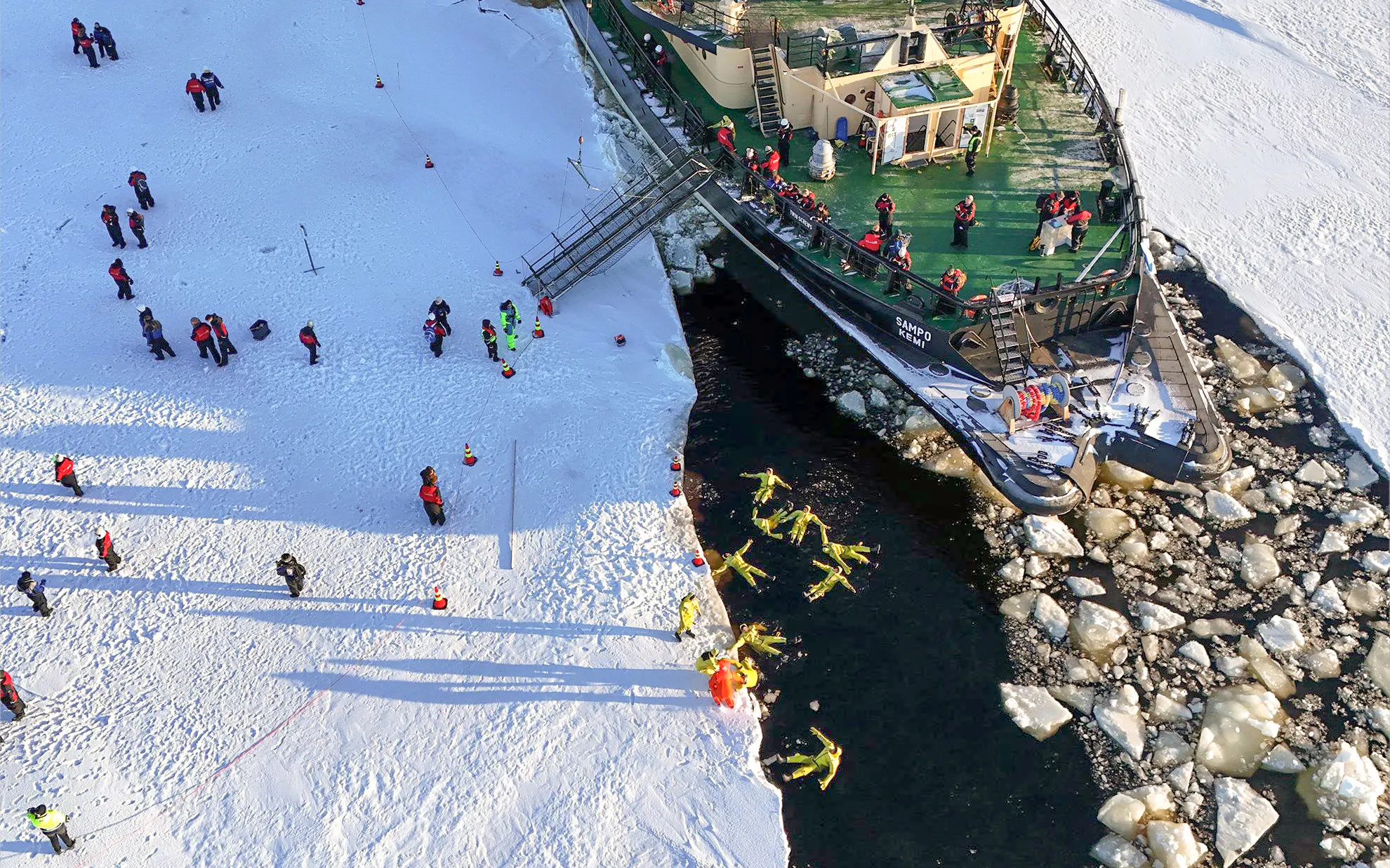Icebreaker Sampo cruise passengers in thermal suits floating in icy water near Kemi, Finland.