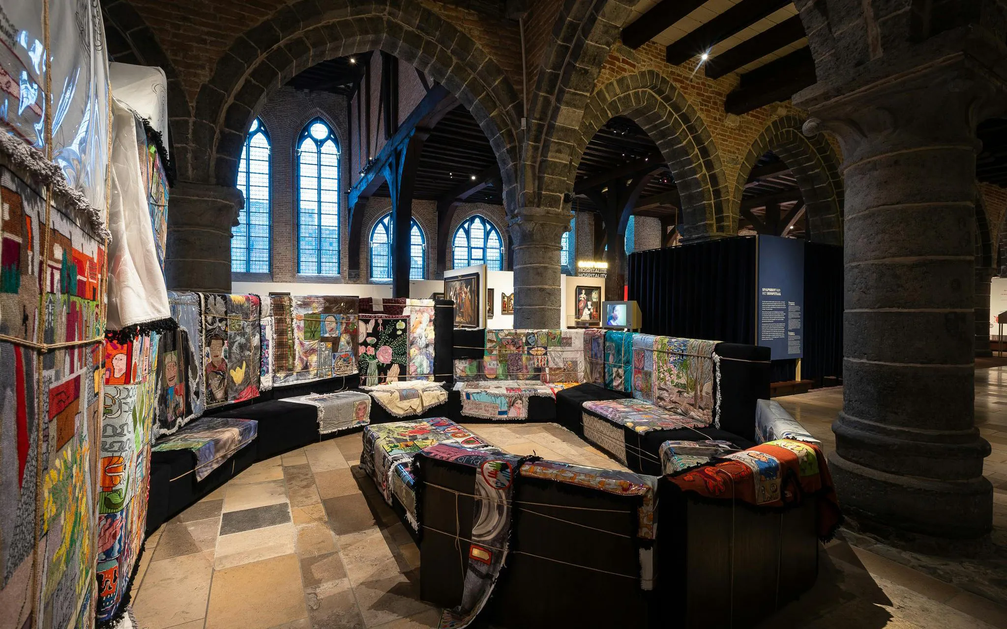 Interior of Museum Sint-Janshospitaal with colorful textile art on benches and stone arches.