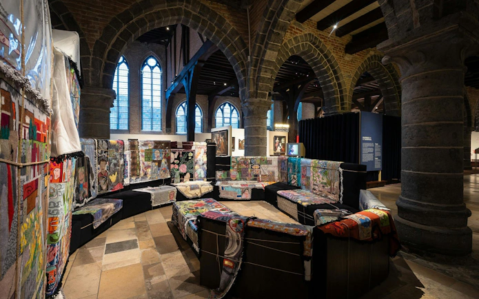 Interior of Museum Sint-Janshospitaal with colorful textile art on benches and stone arches.