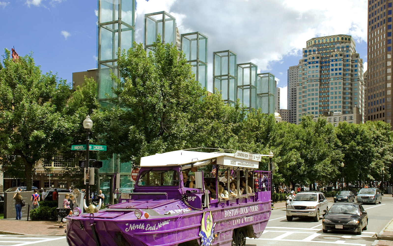 Boston Duck Tours vehicle on city street near glass towers and trees.