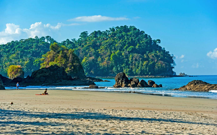 Beach view at Manuel Antonio National Park, Costa Rica, with lush forest and rocky shoreline.