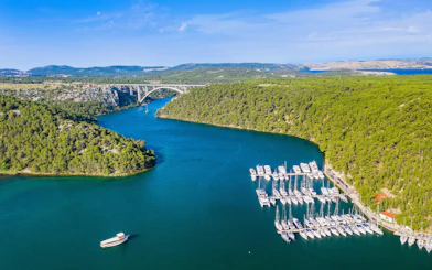 Krka River with boats and marina surrounded by lush greenery and a bridge in the distance.