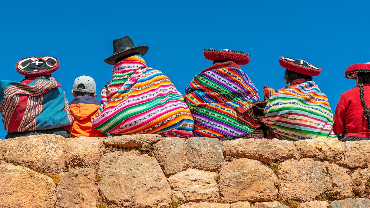 A group of Quechua indigenous women in traditional clothing and a young boy sitting and chatting on an ancient Inca wall