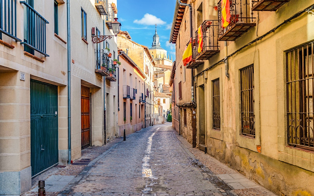 Cobbled medieval street in Segovia with Spanish flags and historic buildings.
