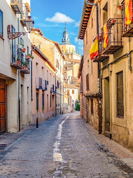 Cobbled medieval street in Segovia with Spanish flags and historic buildings.