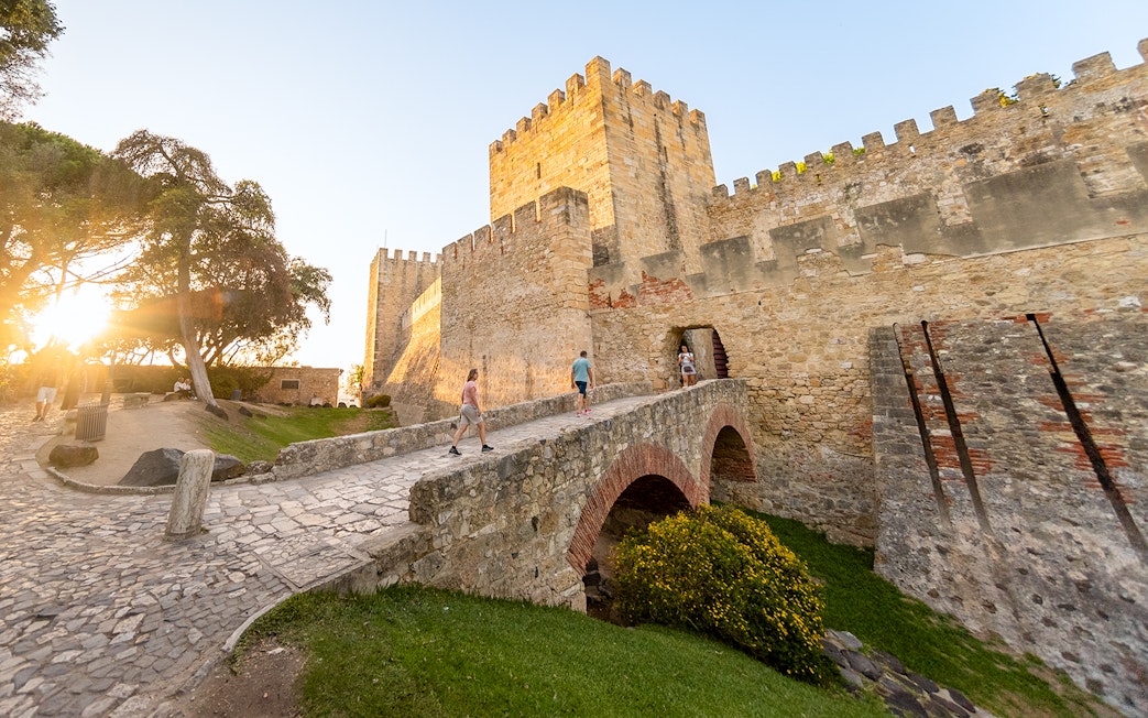 Visitors walking across a stone bridge towards St. George’s Castle entrance in Lisbon.