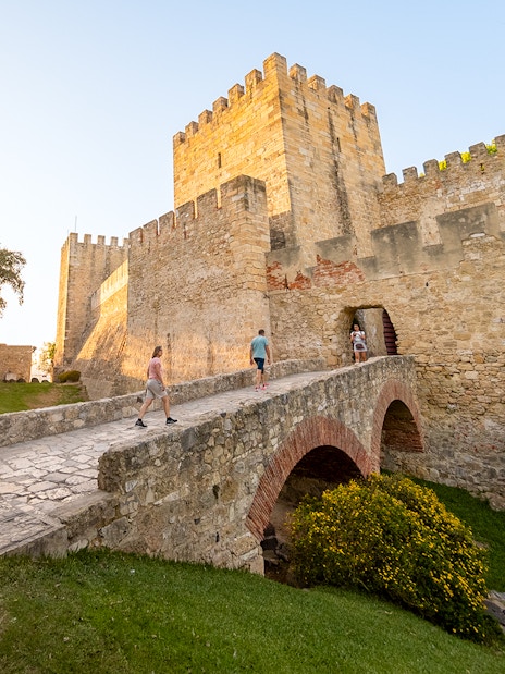 Visitors walking across a stone bridge towards St. George’s Castle entrance in Lisbon.