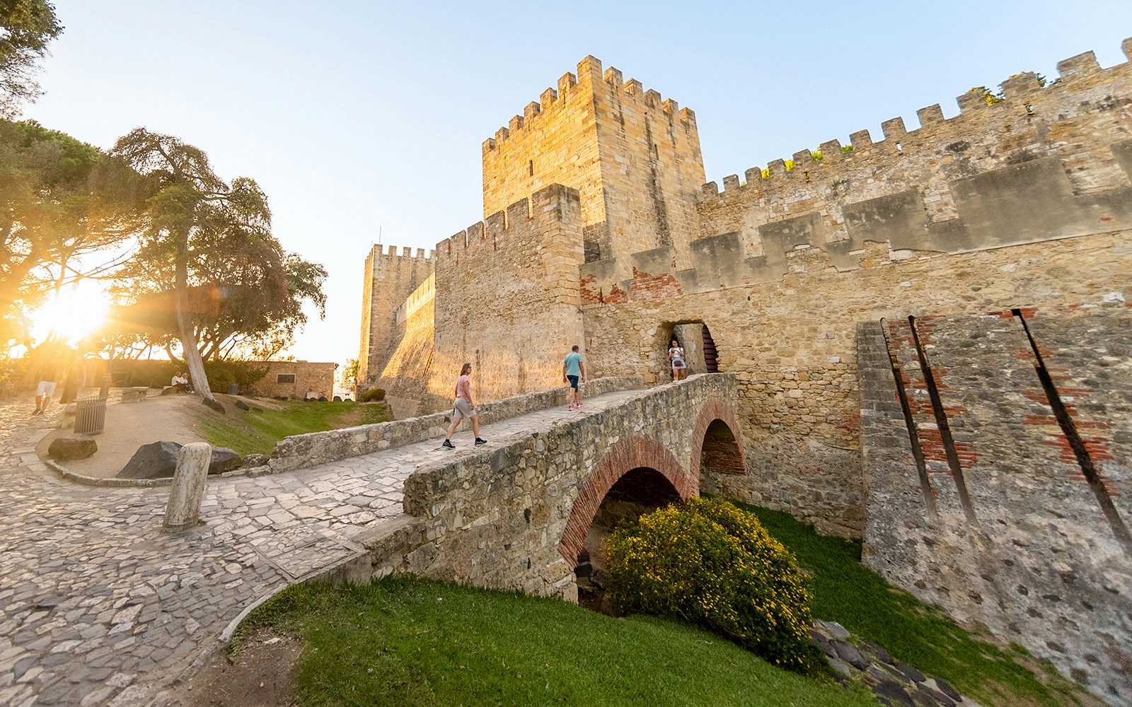 Visitors walking across a stone bridge towards St. George’s Castle entrance in Lisbon.