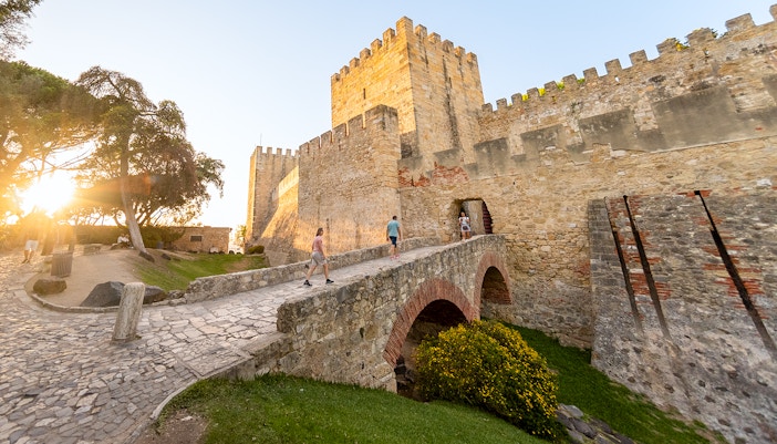 Visitors walking across a stone bridge towards St. George’s Castle entrance in Lisbon.
