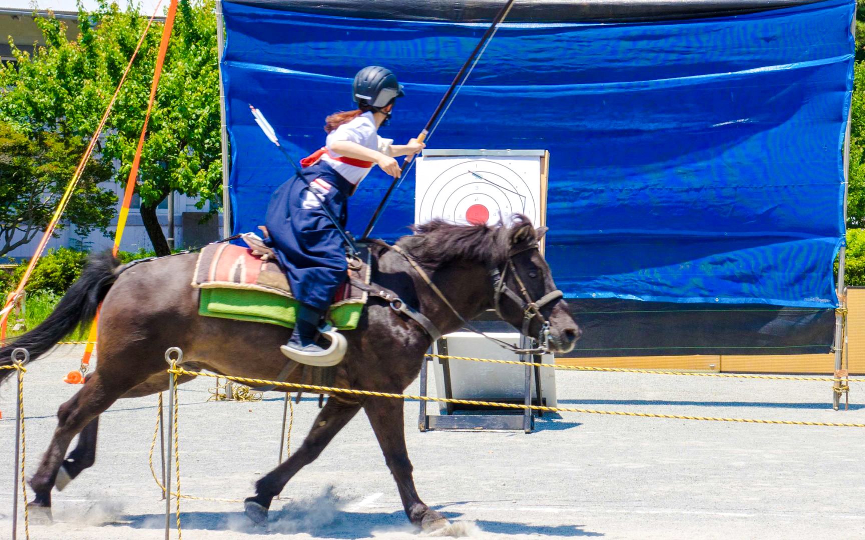 Person practicing Yabusame mounted archery, aiming at a target while riding a horse.