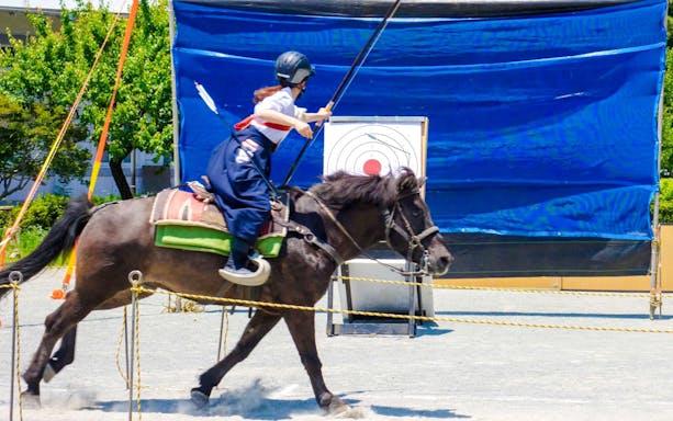 Person practicing Yabusame mounted archery, aiming at a target while riding a horse.
