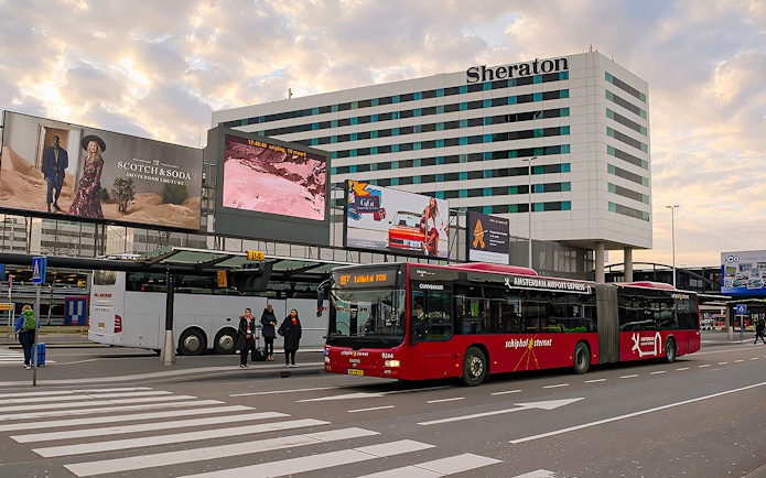 Amsterdam Airport Express bus at Schiphol Airport terminal with Sheraton hotel in background.