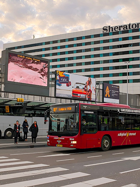 Amsterdam Airport Express bus at Schiphol Airport terminal with Sheraton hotel in background.
