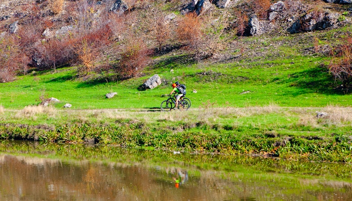 Cyclists riding along a grassy path beside a river in the countryside.