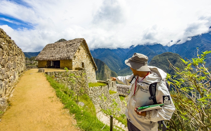 Guide pointing at stone structure in Machu Picchu, Peru, with mountains in the background.
