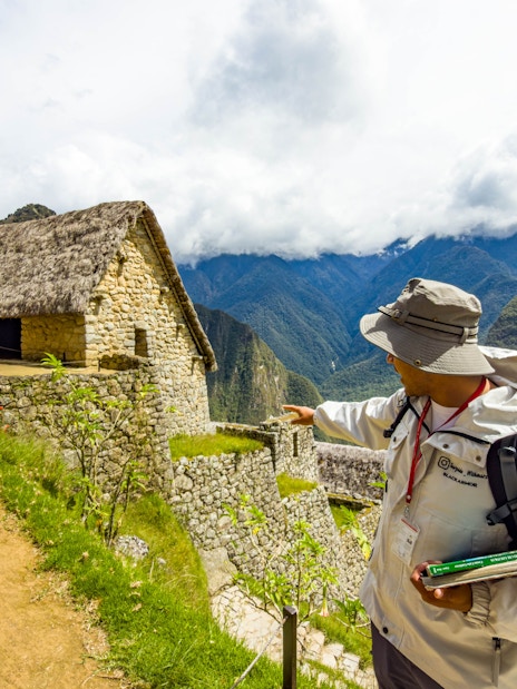 Guide pointing at stone structure in Machu Picchu, Peru, with mountains in the background.