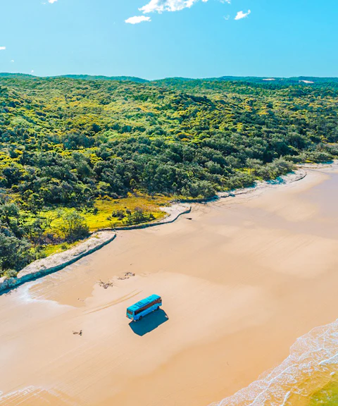 4 wheel drive coach on K'gari beach, Fraser Island, with lush greenery and ocean waves.