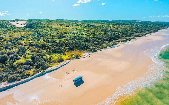 4 wheel drive coach on K'gari beach, Fraser Island, with lush greenery and ocean waves.