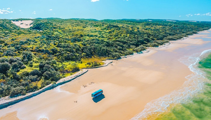 4 wheel drive coach on K'gari beach, Fraser Island, with lush greenery and ocean waves.