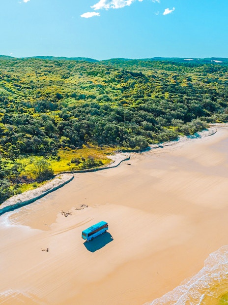 4 wheel drive coach on K'gari beach, Fraser Island, with lush greenery and ocean waves.