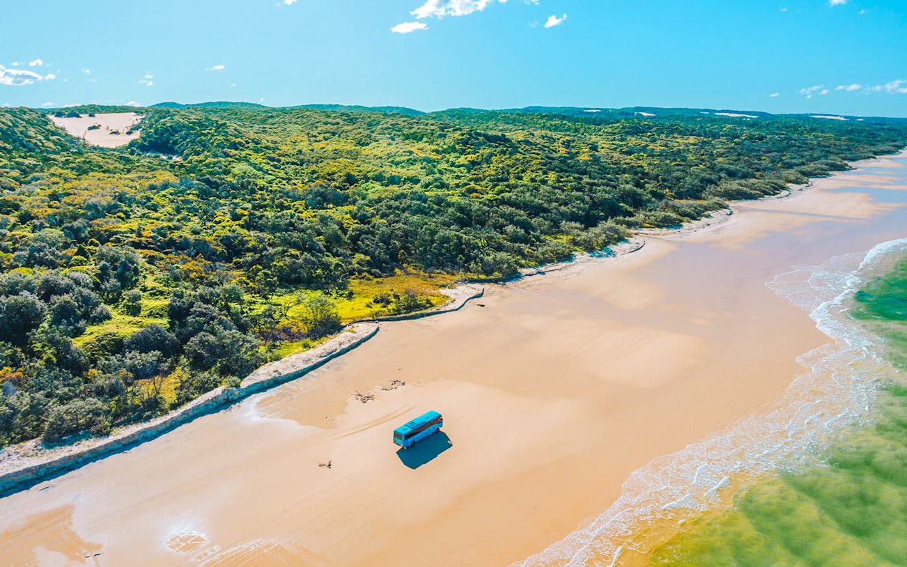 4 wheel drive coach on K'gari beach, Fraser Island, with lush greenery and ocean waves.