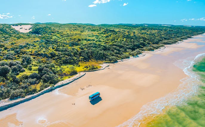4 wheel drive coach on K'gari beach, Fraser Island, with lush greenery and ocean waves.