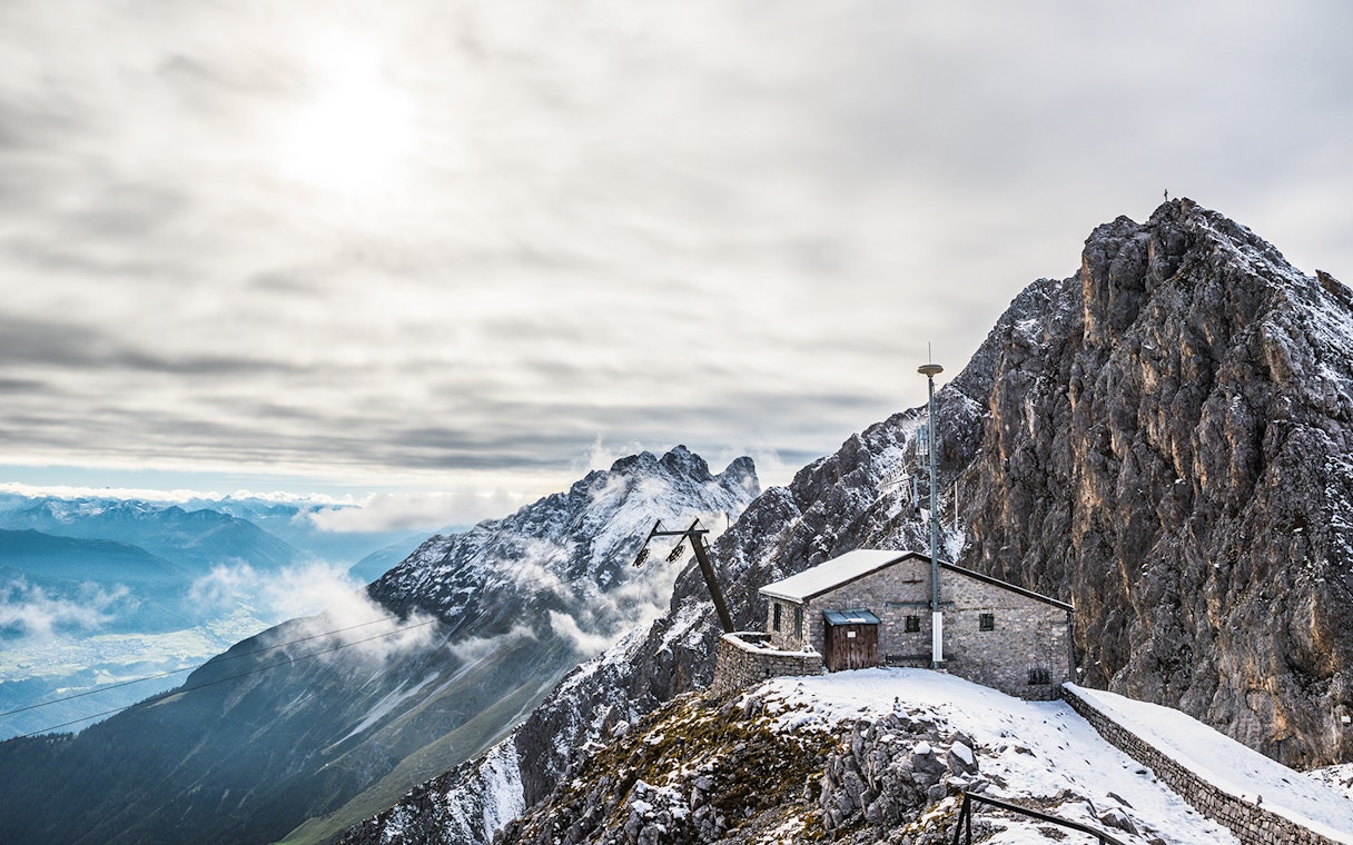 Mountain hut on snowy peak with panoramic view of Alps, clouds, and valley below.
