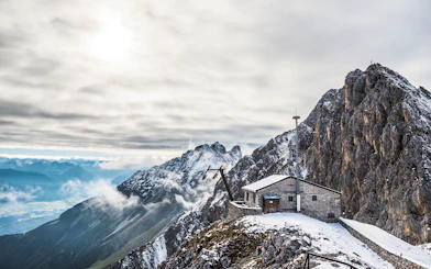 Mountain hut on snowy peak with panoramic view of Alps, clouds, and valley below.