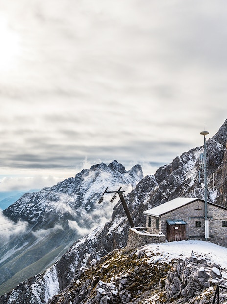 Mountain hut on snowy peak with panoramic view of Alps, clouds, and valley below.