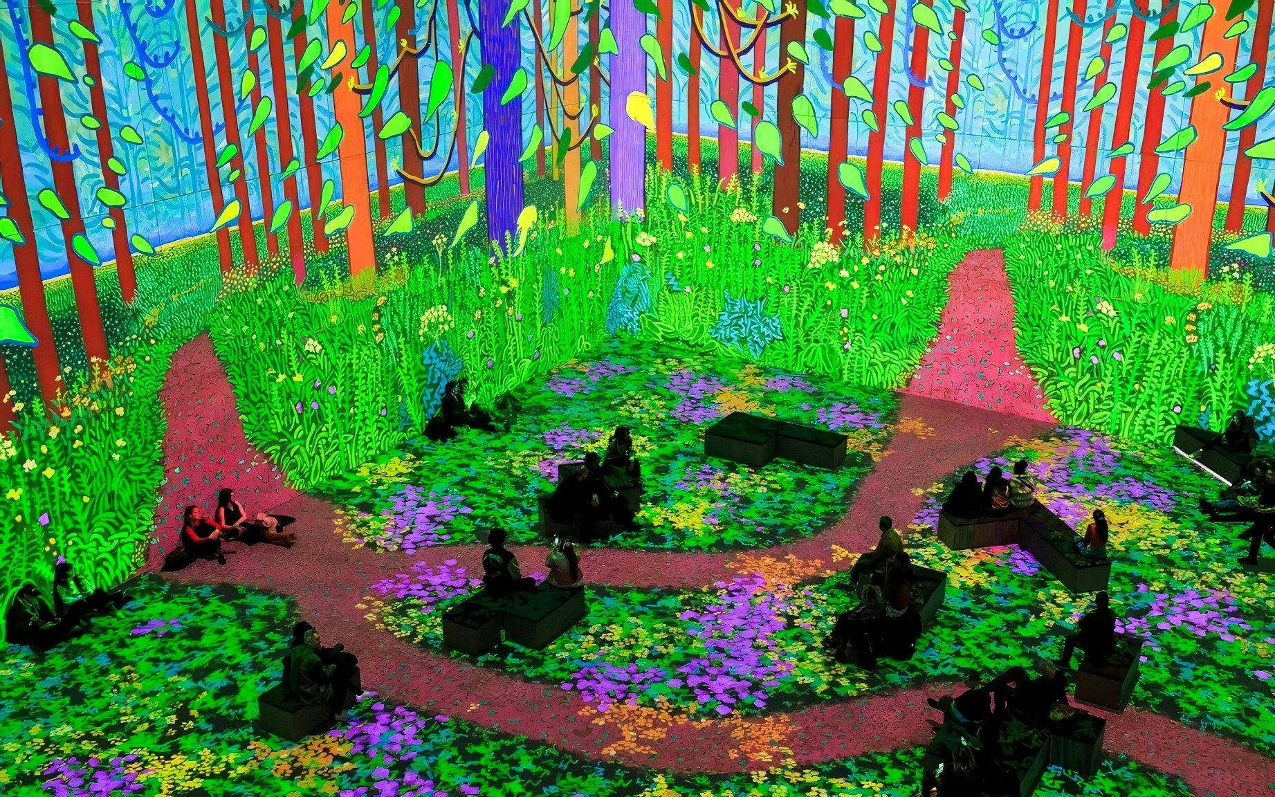 Immersive digital forest scene at David Hockney: Bigger & Closer exhibition, featuring vibrant paths and seated visitors.