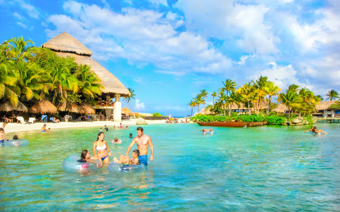 Visitors enjoying the lagoon at Xcaret Park, surrounded by palm trees and thatched-roof structures.