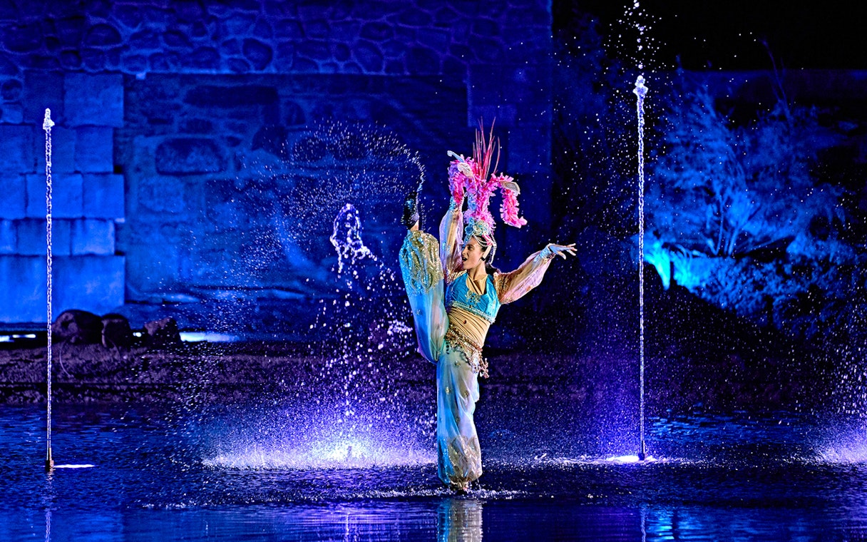 Performer in colorful costume dancing amid water fountains at Puy du Fou España night show.
