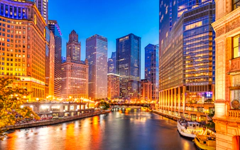 Chicago downtown skyline with illuminated skyscrapers along the Chicago River at dusk.