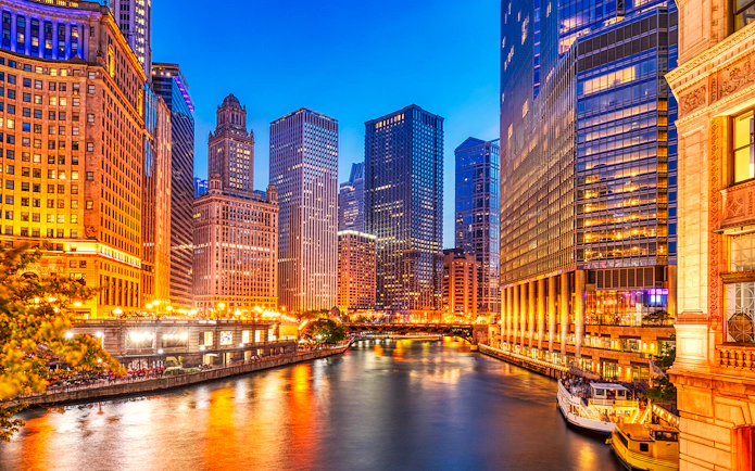Chicago downtown skyline with illuminated skyscrapers along the Chicago River at dusk.