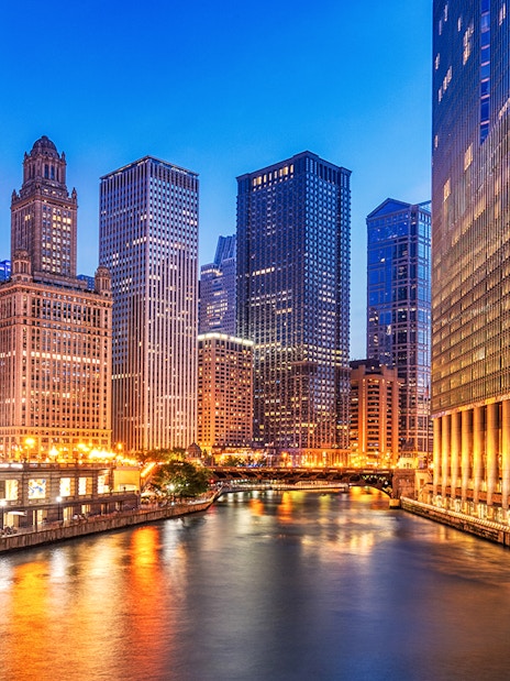 Chicago downtown skyline with illuminated skyscrapers along the Chicago River at dusk.