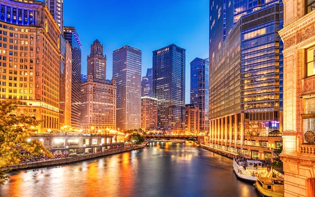 Chicago downtown skyline with illuminated skyscrapers along the Chicago River at dusk.