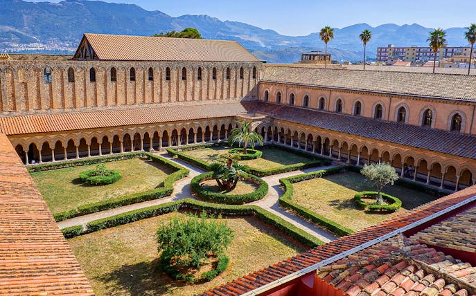 Monreale Cathedral cloister garden with arches and mountain backdrop, Sicily.
