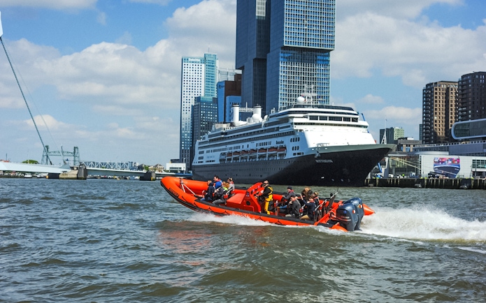 RIB speedboat with tourists in Rotterdam Harbour near cruise ship and skyline.