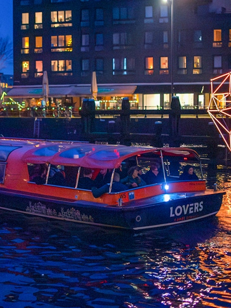 Amsterdam canal boat with passengers on a dinner cruise at night, illuminated by city lights.